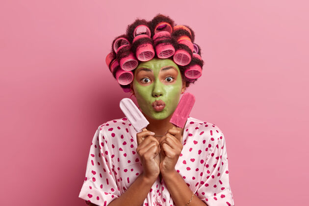 女性漂亮女人的肖像使嘴唇圆润 涂上绿色的面膜 卷发器 夹着两个美味的冰淇淋 穿着丝绸长袍姿势粉色站