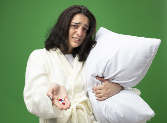 穿恼怒的年轻白种人生病的女孩穿着长袍拿着枕头向相机伸出医疗胶囊看着隔离在绿色背景上的相机枕头绿色长袍