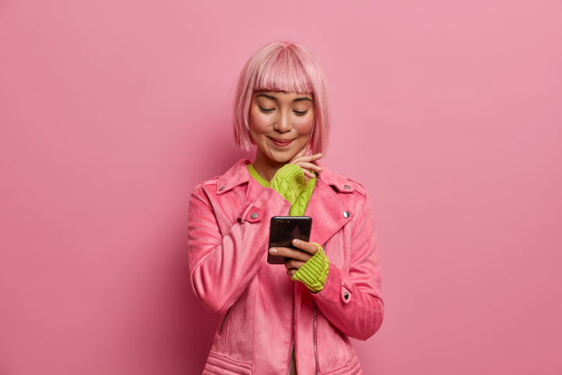 姿势满意迷人的女性社交媒体博客 时尚的发型 手持智能手机 在互联网上阅读文章欢呼亚洲人粉色假发