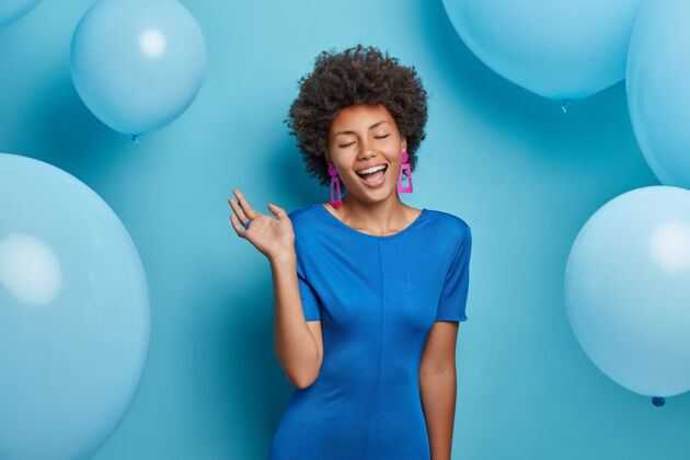 节日喜出望外的美国黑人妇女高兴地闭上眼睛 穿着优雅的蓝色连衣裙 在节日气球上做模特 在庆祝活动中摆姿势 有派对的气氛生日女孩随着音乐的节奏移动女士时尚高兴