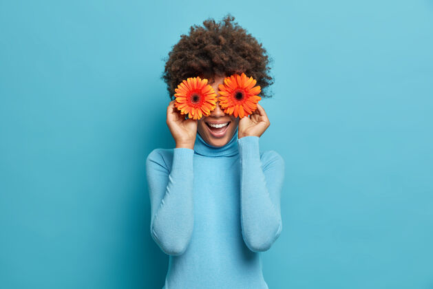 精力充沛顽皮积极的非洲裔美国女人用两只橙色非洲菊遮住眼睛 享受春天的时光 鲜花 开心 穿着随意 心情自然欢笑