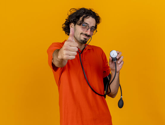 拇指年轻的白种人戴着眼镜 手持听诊器 手持血压计 看着相机 在橙色背景下 用复制空间孤立地竖起大拇指年轻疾病听诊器