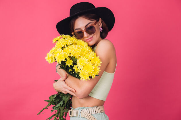 深色戴着帽子和墨镜的时髦女人 拥抱着一大束黄紫苑 春意盎然 淡定微笑的孤立空间祝贺植物学菊花