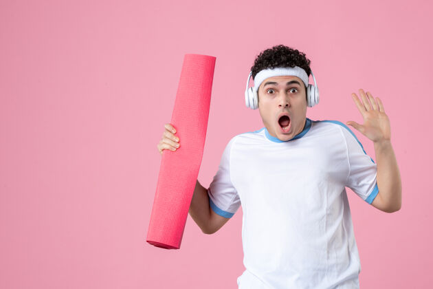 前面正面图：穿着运动服的年轻男性 粉色墙上有瑜伽垫和耳机耳机惊喜运动