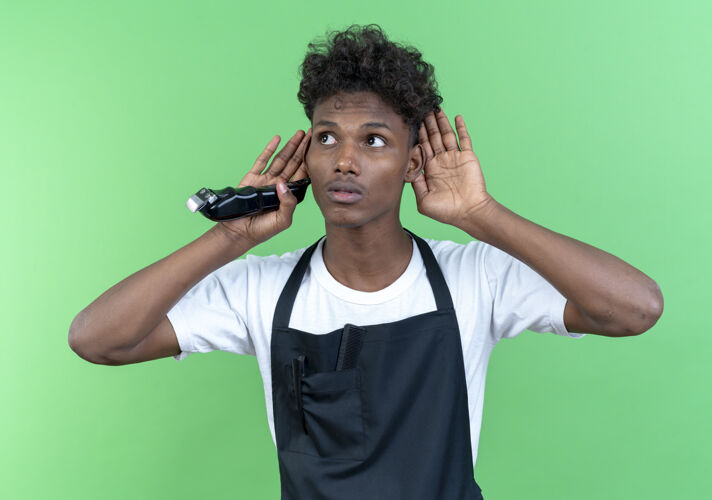 思考年轻的美国黑人男理发师穿着制服 拿着理发器 在绿色的墙上展示着孤立的倾听姿态手势显示壁板