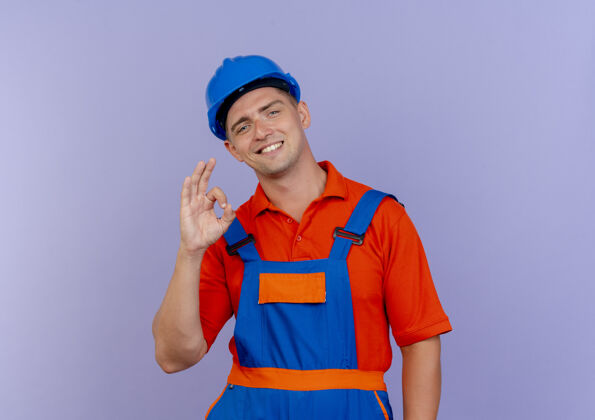 手势微笑的年轻男性建筑工人穿着制服 戴着安全帽 在紫色的地板上显示出良好的姿态紫色制服建筑工人