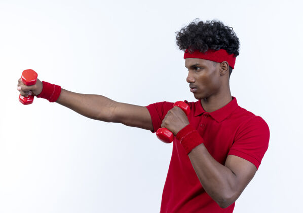 年轻站在侧视图年轻的黑人运动男子戴着头带和手环举着哑铃隔离在白色哑铃侧写手势