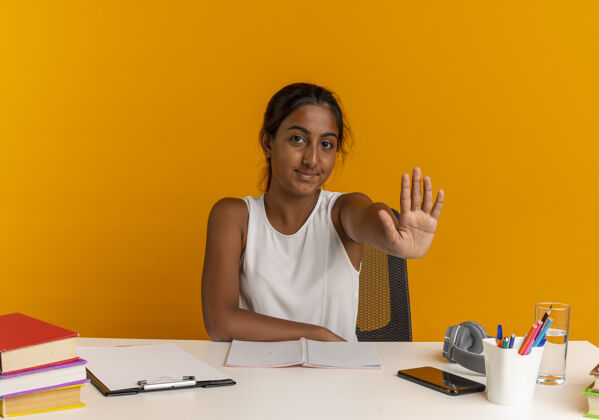 女学生坐在书桌旁的小女孩很高兴 手里拿着学习用具 在橘子上做着停车的手势请手势书桌