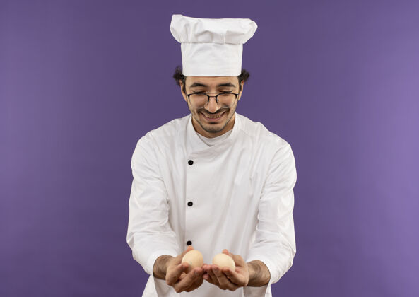 紫色高兴的年轻男厨师穿着厨师制服 戴着眼镜 拿着紫色的鸡蛋看着背景年轻厨师