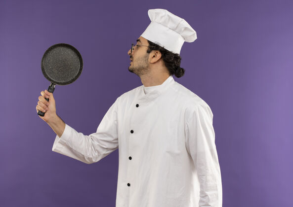 视图站在剖面图年轻的男厨师穿着厨师制服和眼镜拿着和看着紫色的煎锅紫色眼镜持有