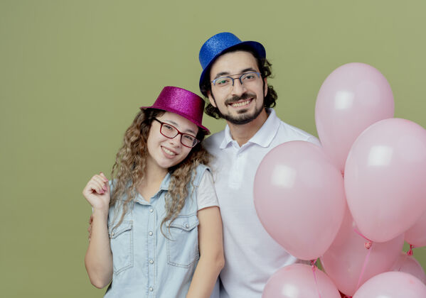 帽子微笑着的年轻夫妇戴着粉色和蓝色的帽子站在橄榄绿上孤立的气球旁情侣粉色站立