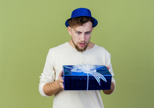 表情愁眉不展的年轻帅哥戴着派对帽 拿着礼物盒看着橄榄绿的墙上孤立的礼物盒人抱着皱眉