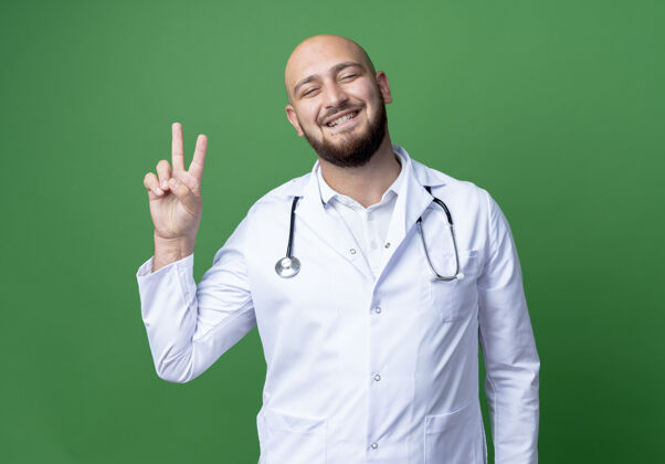 年轻微笑着的年轻男医生穿着医用长袍和听诊器 在绿色上显示出良好的姿势听诊器微笑医疗