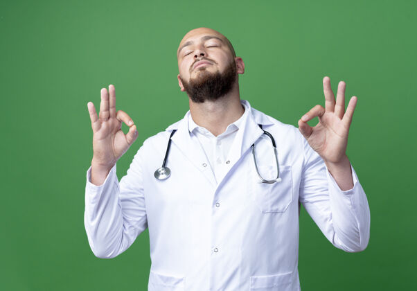 听诊器年轻的男医生戴着医用长袍和听诊器 闭着眼睛 在绿色的地面上显示出很好的手势年轻绿色医生