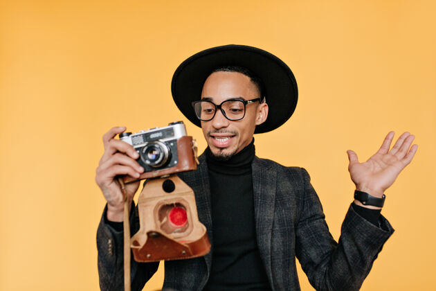 前景一个戴着帽子和西装的非洲男人拿着相机 表示惊讶无忧无虑的黑人在黄色墙上拍照时的肖像非洲人服装积极