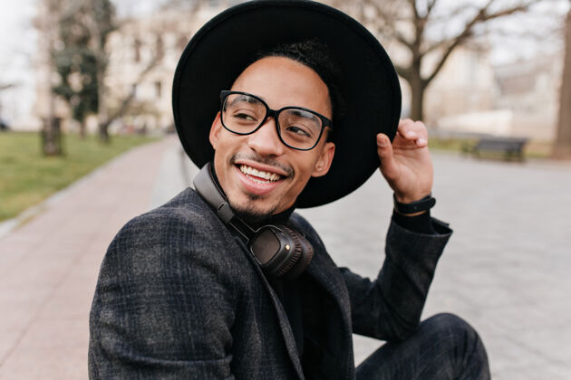 快乐穿着羊毛夹克的快乐的非洲男人坐在城市的户外肖像欣喜若狂的黑人摸着帽子微笑着看向别处的照片帅气休闲年轻