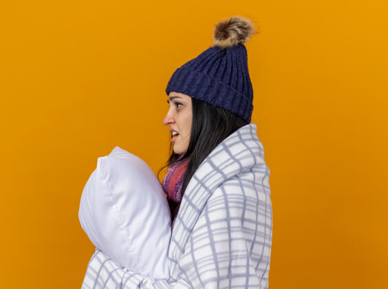 侧写印象深刻的年轻白种人生病的女孩戴着冬天的帽子和围巾包裹在格子布站在纵断面图抱着枕头看直孤立的橙色背景与复制空间复制帽子包