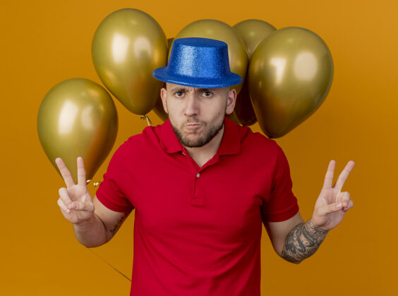 气球皱眉的年轻帅哥戴着派对帽站在气球前看着前面做着和平手势孤立在橙色的墙上英俊立场帽子