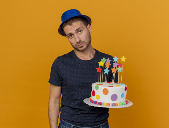 穿着失望的帅哥戴着蓝色的派对帽 把生日蛋糕孤立地放在橙色的墙上人表情生日