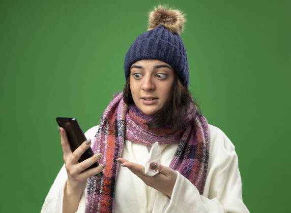 年轻人印象深刻的年轻生病的女人穿着长袍 戴着冬天的帽子和围巾 拿着手机看着它 手里拿着餐巾隔离在绿色的墙上电话人表情