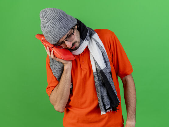 姿势愁眉不展的年轻病人戴着眼镜 帽子和围巾 把热水袋放在脸上 闭着眼睛隔离在绿色的墙上人人包