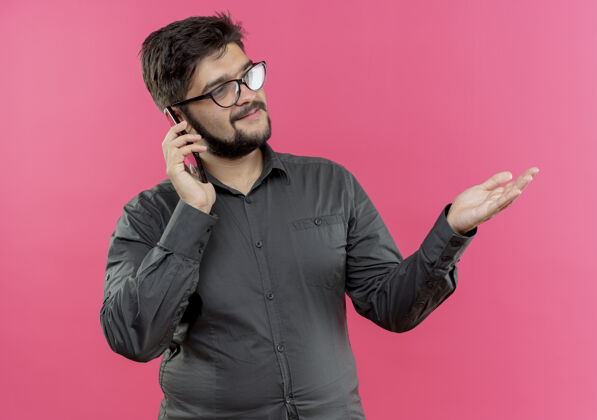 电话看着一旁高兴的戴眼镜的年轻商人讲电话 用手指着隔离在粉红色墙上的一边年轻人穿商务人士