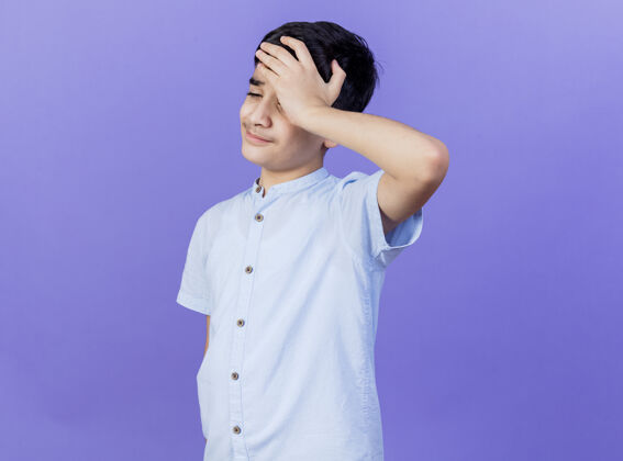 人疼痛的小男孩手放在头上看着紫色的墙上孤立的头痛表情疼痛年轻