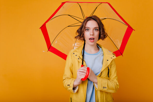 肖像带着惊奇表情的白人女孩站在黄色的墙上 手里拿着一把红色的阳伞这是一张梦幻般的黑发女士穿着秋装 打着时髦的雨伞的照片情绪年轻伞