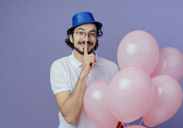 帽子笑容可掬的帅哥戴着眼镜 戴着蓝色的帽子 举着气球 表现出沉默的姿态 孤立在紫色上紫色帅气沉默