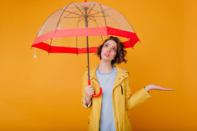 心烦穿着秋雨衣心烦意乱的年轻女模特的室内写真悲伤的卷发女士站在时髦的雨伞下仰望的照片天气年轻下雨