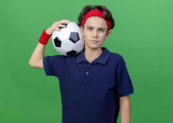 球自信的年轻帅气的运动男孩戴着头带和护腕 戴着牙套 肩上扛着足球 看着绿色墙壁上孤立的前方腕带背带自信