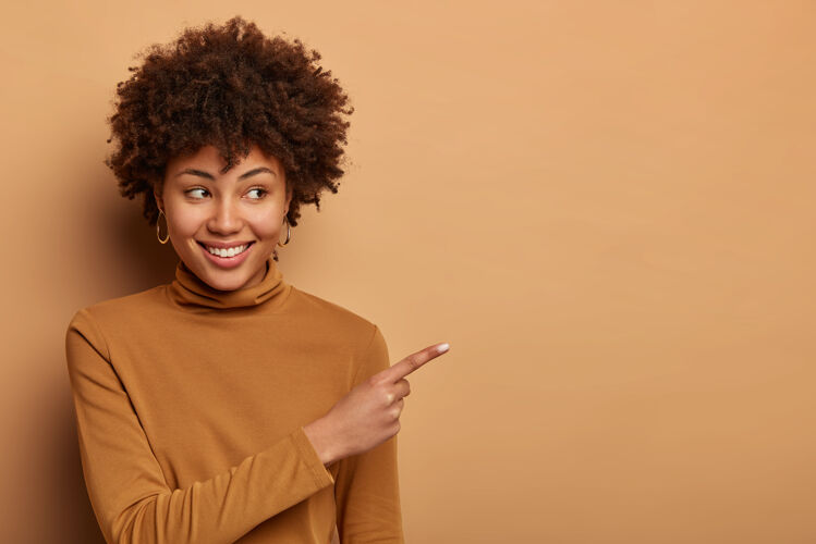 高兴快乐快乐的美国黑人妇女在一旁表示 推销商品 高兴地微笑 穿着棕色高领毛衣 对着棕色墙壁摆姿势广告概念这东西不错看看那里检查女性青少年