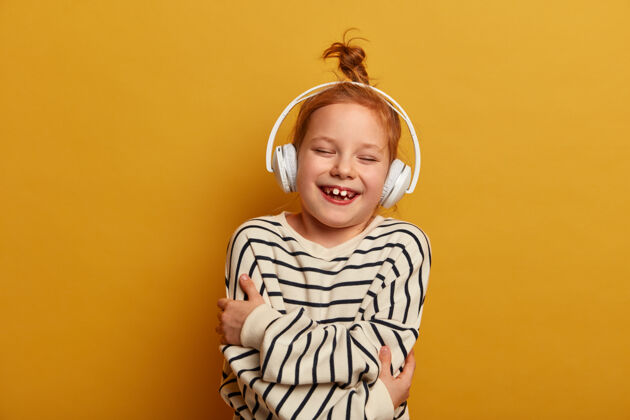 乐观小姜女孩抱着自己 笑着玩着 戴着立体声耳机听音乐 穿条纹套头衫 在黄色的墙上摆姿势 把空闲时间花在最喜欢的爱好上 觉得很有趣耳机高兴心情