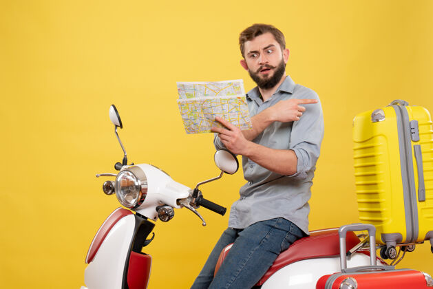 拿着旅游概念的前视图 困惑的年轻人坐在摩托车上 手提箱指向后面 黄色的地图地图背车
