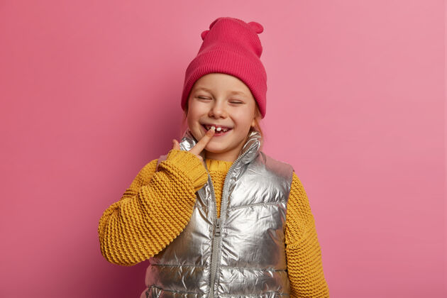 牙齿可爱的小女儿 小女孩儿指着她的新牙 笑容满面 戴帽子 穿毛衣和背心 防龋 关心牙齿 粉彩玫瑰墙上的模特休闲指向孩子