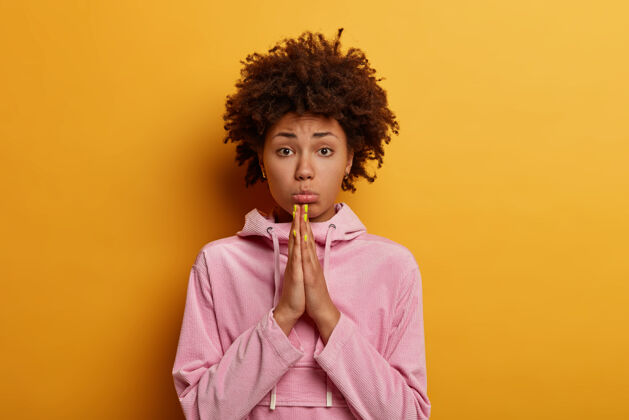 女性不满意的非洲裔美国妇女的照片保持手掌压在一起 祈祷或恳求 乞求您的帮助 钱包嘴唇 看起来悲伤 对黄色的墙壁姿势 穿着休闲的玫瑰色连帽衫情感失望非洲人