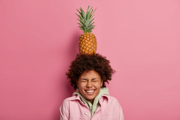 平衡美丽俏皮的女人 非洲式发型 头上有异国情调的菠萝 眯着眼睛 有着牙齿般的微笑 穿着连帽衫 对着粉色粉彩的墙壁摆姿势女人玩着成熟的水果斜视异国情调人