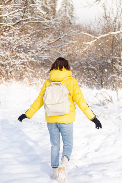 时髦一个穿着亮黄色夹克和牛仔裤的女人背着一个背包在雪景森林里走过雪堆下雪霜冻天气