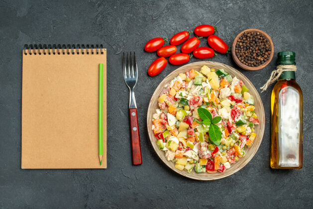 叉子一碗蔬菜沙拉的顶视图 在深灰色背景上 有叉子 油瓶 西红柿 胡椒和记事本蔬菜壁板背景