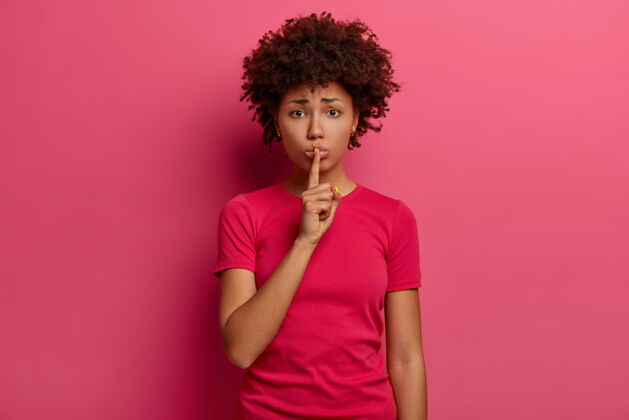 发型请闭嘴失望的年轻非洲裔美国女人做了一个安静的手势 表示沉默 皱眉 把食指按在嘴唇上 穿着休闲t恤 在粉红色的墙上摆姿势女孩手势站立