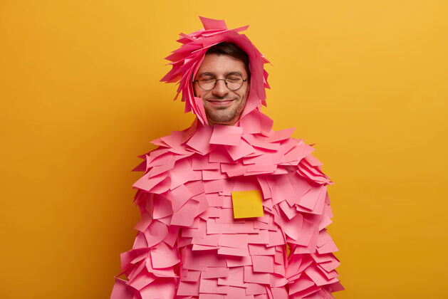 笔记积极的家伙在身体和头部贴上粉红色的胶纸 用胶纸做创意服装 戴眼镜 在办公室工作 隔离在黄色的墙上 闭上眼睛胡茬姿势眼镜