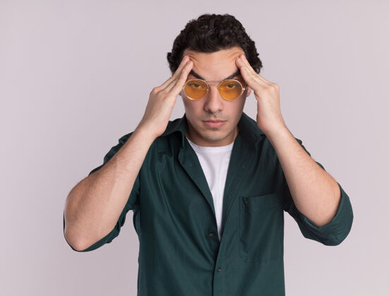 衬衫一个穿绿衬衫戴眼镜的年轻人站在白墙上摸着头累得头痛男人头疲倦