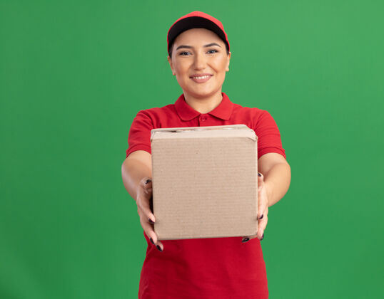 展示身着红色制服 头戴礼帽的快乐的年轻女送货员站在绿色的墙上 面带微笑地看着前面的纸板箱女人制服站立