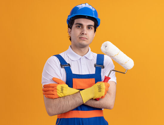 年轻愤怒的年轻建筑工人身穿施工制服 戴着安全帽 戴着橡胶手套 拿着油漆辊 站在橙色的墙上 神情严肃自信地看着前方建设者橡胶滚轴