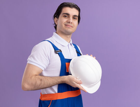 站立身穿建筑制服的年轻建筑工人手持安全帽 自信地站在紫色的墙上 望着前方建设者安全自信