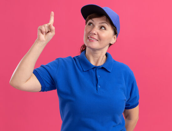 女人身穿蓝色制服 头戴鸭舌帽的中年女送货员脸上挂着微笑 用食指指着粉色墙上站着的东西脸指向手指