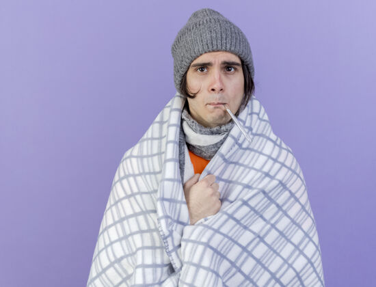 拿着看着镜头虚弱的年轻病夫戴着冬天的帽子 围巾裹着格子布 嘴里拿着温度计 隔离在紫色的背景上嘴包裹年轻