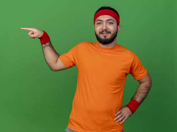 腕带微笑的年轻运动男子戴着头带和腕带点在一边 把手放在臀部隔离在绿色背景与复制空间运动年轻壁板