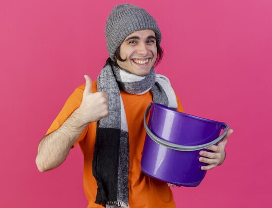 微笑带着微笑的年轻病人戴着冬天的帽子 戴着围巾 拿着塑料桶 在粉色背景上孤立地竖起大拇指围巾塑料拇指