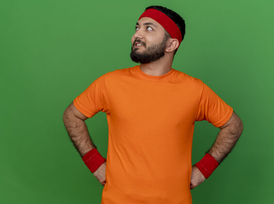 头带面带微笑的年轻运动型男子 戴着头带和腕带 手放在臀部 与绿色背景隔离壁板运动手